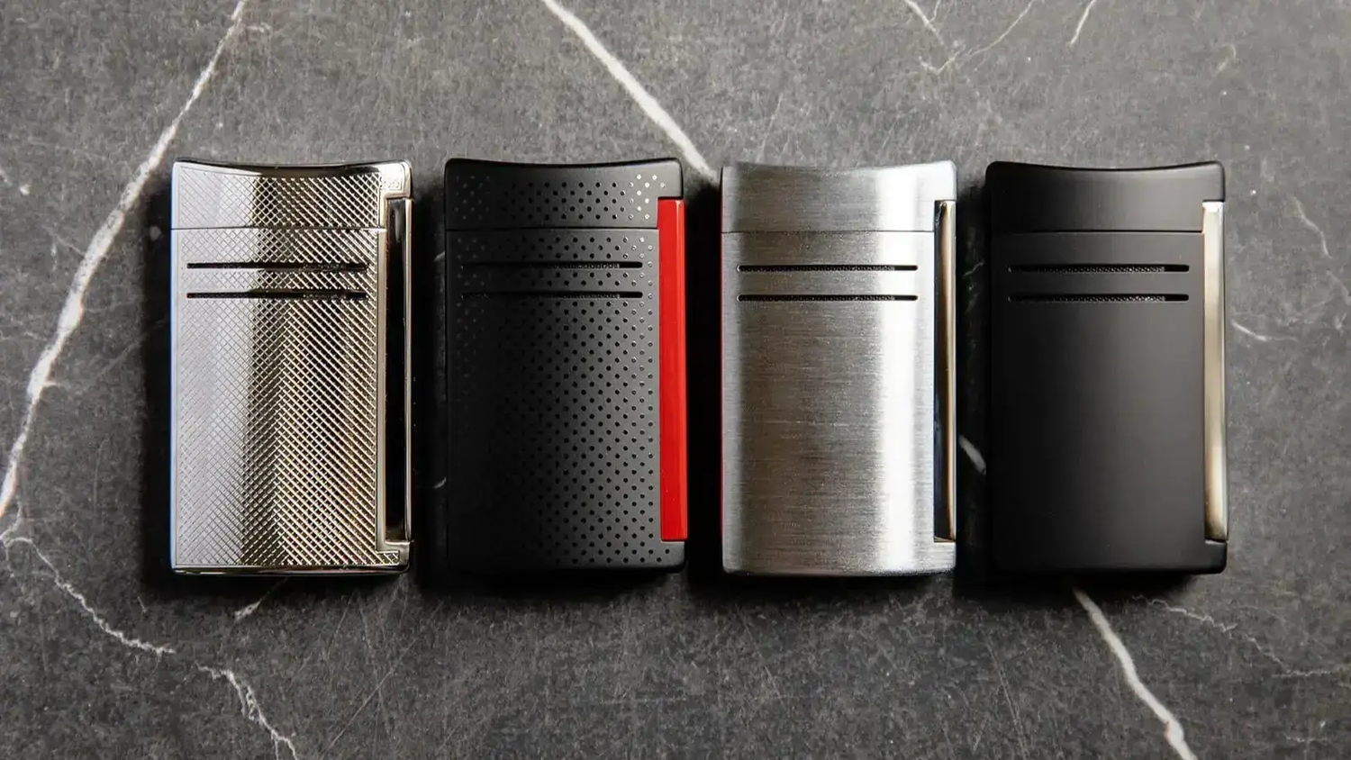S.T. Dupont Maxijet torch lighters on marble background. Left to right - chrome grid, punched red and black, chrome, matte black